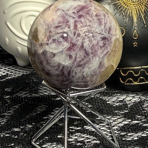 May include: A polished crystal sphere with purple and white veining, displayed on a silver metal stand. The sphere has a translucent quality, and the stand has a geometric design. The background includes a black lace-like fabric and decorative objects.