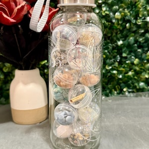 May include: A clear glass water bottle filled with small, clear plastic spheres containing various decorative elements. The bottle has a silver lid and a white strap. The bottle is decorated with etched designs. The spheres contain items like small stones and paper scrolls.