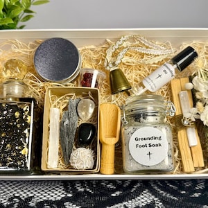 May include: A white box filled with various spiritual items. Includes a jar of foot soak, grounding oil, crystals, a bell, a wooden scoop, and a decorative bottle. The box is filled with straw.