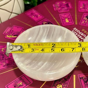 Może przedstawiać: Biała miska z selenitu o gładkiej, falistej powierzchni. Miska ma średnicę około 7,6 cm i jest mierzona ż&oacute;łtą miarką.