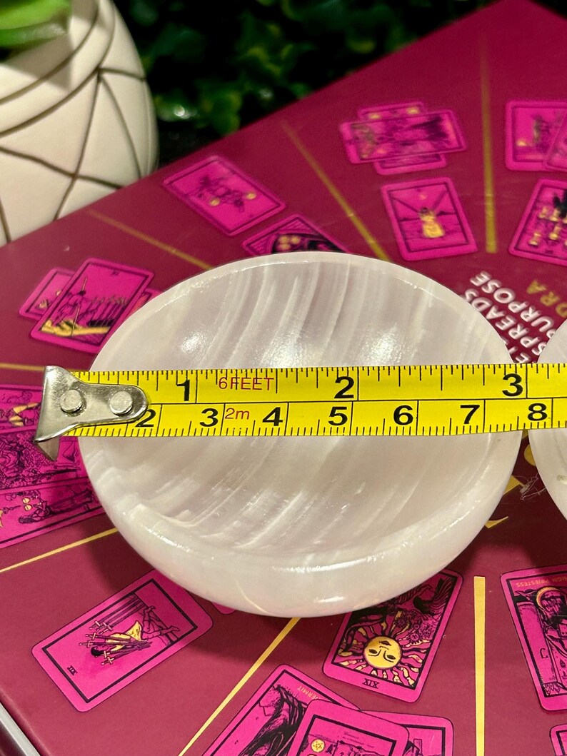 Może przedstawiać: Biała miska z selenitu o gładkiej, falistej powierzchni. Miska ma średnicę około 7,6 cm i jest mierzona ż&oacute;łtą miarką.