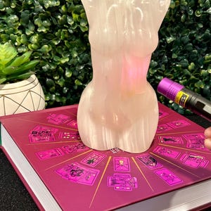 May include: A white selenite sculpture in the shape of a human torso, sitting on top of a book with a pink cover and a tarot card design. The book is titled "The Wild Unknown Tarot".