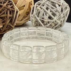 May include: A clear quartz bracelet with rectangular beads. The bracelet is made of translucent, polished quartz and is displayed on a white surface. Decorative woven spheres are in the background.