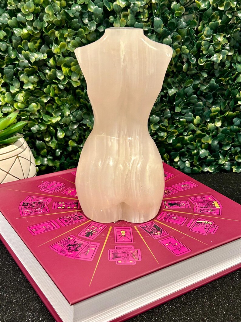 May include: A white, smooth, polished,  selenite torso sculpture. The sculpture is sitting on a book with a pink cover and a circular design of tarot cards.