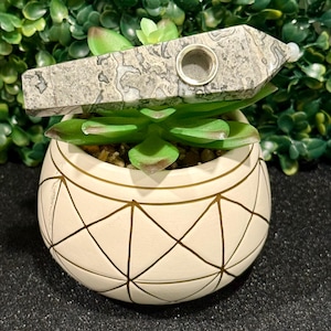 May include: A gray and white stone pipe with a round hole in the center. The pipe is sitting on top of a white planter with a geometric pattern.