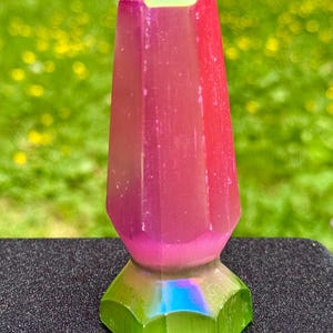 May include: A vibrant, iridescent crystal point in shades of pink, red, and green. The crystal has a faceted base and a pointed top. The colors shift with the light, creating a rainbow effect. The crystal is displayed on a dark surface with a blurred green background.