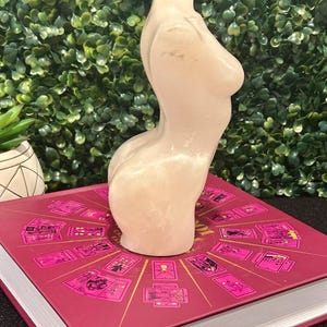 May include: A white, glittery, resin sculpture of a torso. The sculpture is sitting on a book with a pink cover and a design of tarot cards.