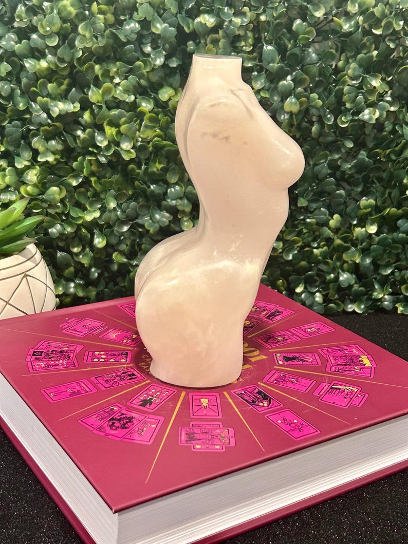 May include: A white, glittery, resin sculpture of a torso. The sculpture is sitting on a book with a pink cover and a design of tarot cards.