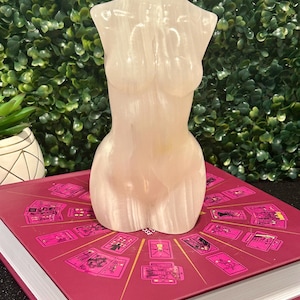 May include: A white selenite crystal carved in the shape of a human torso. The crystal is sitting on a book with a pink cover and a design of tarot cards.