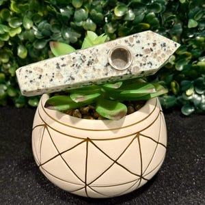 May include: A white and gray speckled stone pipe with a round hole for smoking. The pipe is sitting on top of a white planter with a geometric pattern.