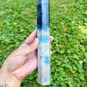 May include: A multi-colored crystal obelisk with a pointed top. The crystal features bands of dark blue, light blue, and beige. The obelisk is held against a backdrop of green grass.