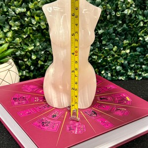 May include: A white, translucent, polished selenite crystal sculpture in the shape of a human torso. The sculpture is approximately 7 inches tall and is sitting on a book with a pink cover.