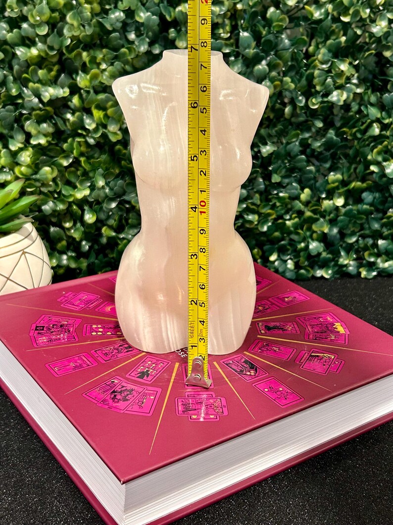 May include: A white, translucent, polished selenite crystal sculpture in the shape of a human torso. The sculpture is approximately 7 inches tall and is sitting on a book with a pink cover.