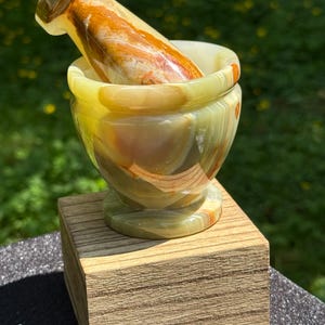 May include: A handcrafted mortar and pestle set made of green and orange onyx stone. The mortar is bowl-shaped, and the pestle is cylindrical. The set is displayed on a small wooden block, perfect for grinding herbs and spices.