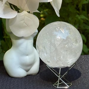 May include: A clear quartz crystal sphere, approximately 4 inches in diameter, sits on a silver metal stand. A white ceramic vase in the shape of a torso is to the left, with white floral accents above. The background is a blurred green.