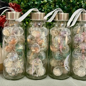 May include: Four clear glass bottles filled with small, clear plastic spheres containing various decorative elements. Each bottle has a silver lid and a gray strap. The bottles are decorated with stickers and filled with colorful objects.