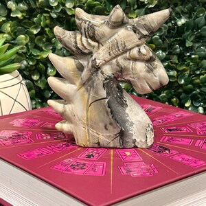 May include: A carved stone unicorn figurine with a white, gray, and black striped pattern. The figurine is sitting on a red book with a pink and gold design.