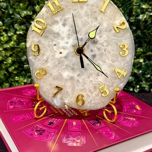 May include: White agate clock with gold-colored numbers and hands, sitting on a gold-colored stand. The clock is on top of a book with a pink cover and gold-colored text.