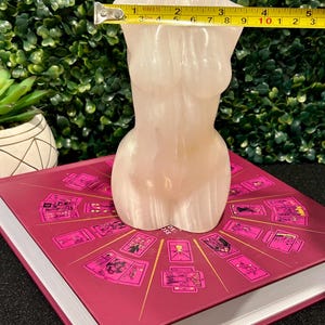 May include: A white, translucent, sculpted female torso figurine, approximately 10 inches tall, sitting on a book with a pink cover and a design of tarot cards.