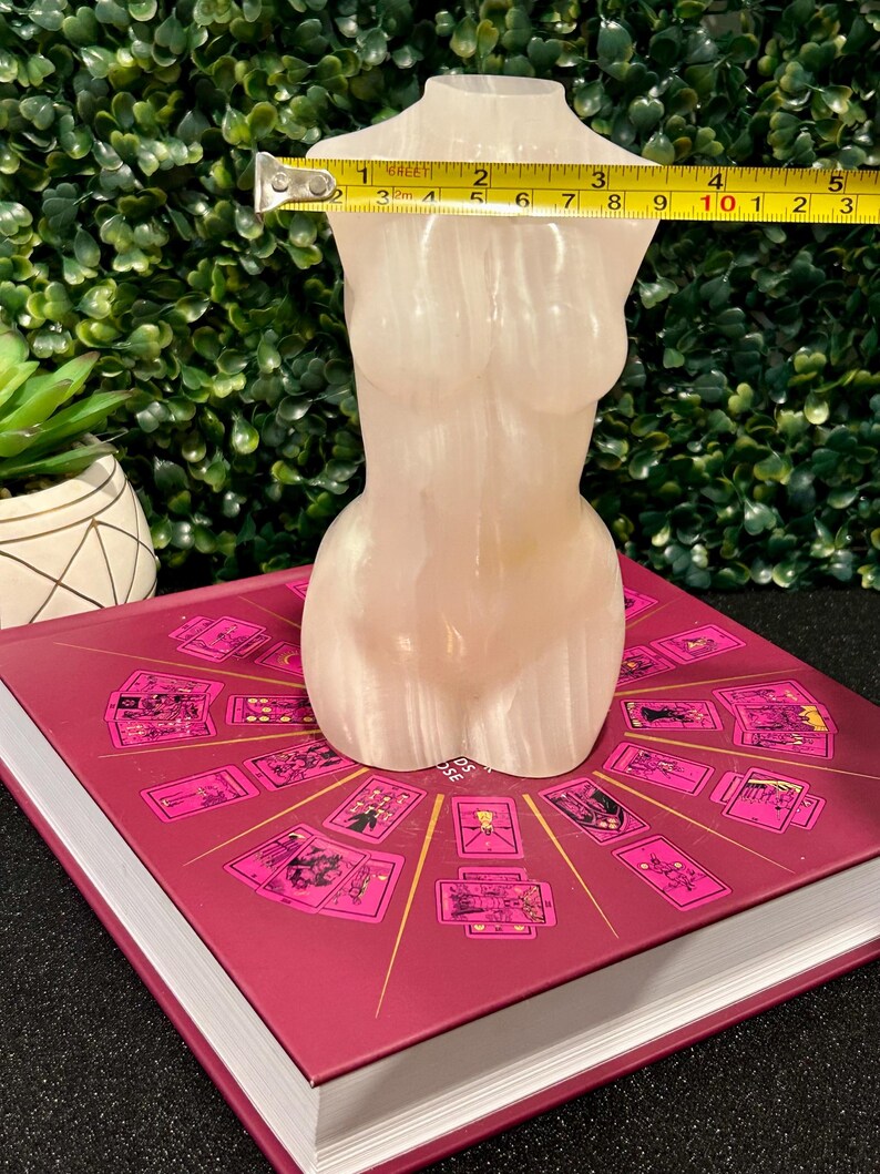 May include: A white, translucent, sculpted female torso figurine, approximately 10 inches tall, sitting on a book with a pink cover and a design of tarot cards.
