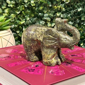 May include: Green and brown speckled stone elephant figurine. The elephant is standing on a book with a pink cover and a design of playing cards.