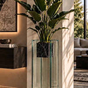 Puede incluir: Una maceta de suelo alta y cuadrada de cristal transparente con un cubo interior de patrón de mármol negro. Una gran planta verde de hojas anchas y alargadas crece desde el centro de la maceta, extendiéndose hacia arriba con follaje vivo.