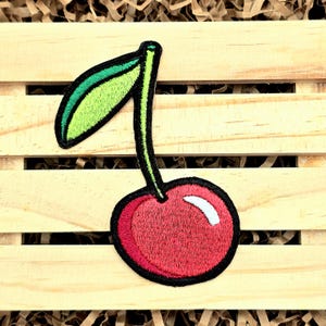 May include: A red and green embroidered cherry patch with a white highlight on the cherry. The cherry has a green stem and leaf.