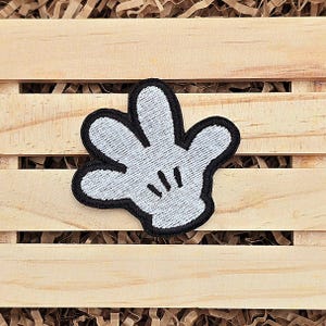 May include: A white felt patch with a black outline of a cartoon hand with four fingers. The hand is in a waving gesture.