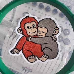May include: Embroidered patch featuring two cartoon monkeys hugging. One monkey is red-orange, the other is gray. The patch has a white border and is set against a textured background, partially framed by a green hoop.