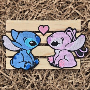 May include: Embroidered patches featuring the characters Stitch and Angel from Lilo & Stitch, facing each other with a pink heart above. Stitch is blue, Angel is pink, and both have black outlines. The patches are on a wooden slat background.