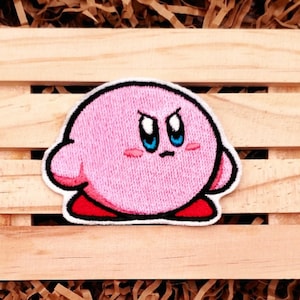 May include: Pink and white embroidered patch of a cartoon character with blue eyes and a frowning expression. The character is pink and has a red and white outline.