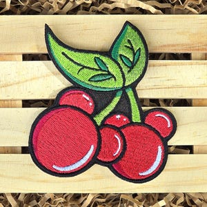 May include: Embroidered patch featuring a cluster of red cherries with white highlights and a green leaf. The patch has a black border and is set against a wooden background.