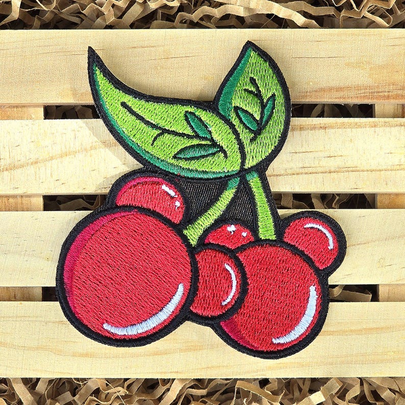May include: Embroidered patch of a cluster of red cherries with white highlights and a green leaf. The patch has a black border, set against a wooden background.