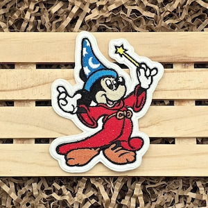 May include: Embroidered patch featuring a cartoon mouse in a red robe and blue wizard hat, holding a yellow star-tipped wand. The mouse is smiling, with black and white details. The patch is outlined in white.