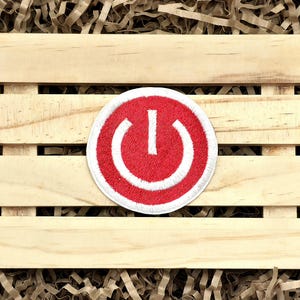 May include: A round, red and white embroidered patch featuring a power button symbol. The patch has a white border and is set against a wooden crate with brown paper filler.