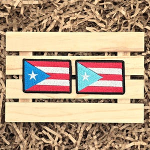 May include: Two embroidered patches featuring the flag of Puerto Rico. Each patch has a blue triangle with a white star, red and white stripes, and a black border. The patches are displayed on a wooden pallet.