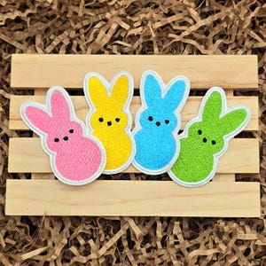 May include: Four embroidered iron-on patches shaped like bunnies in pink, yellow, blue, and green. The bunnies have white outlines and black eyes.