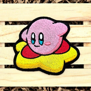 May include: Embroidered patch featuring a pink character with large blue eyes, a red collar, and a yellow base. The patch has a black border and is likely for clothing or accessories.