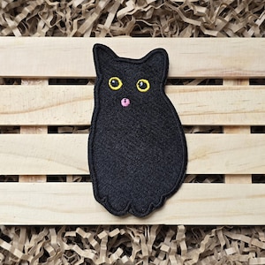 May include: A black cat patch with yellow eyes and a pink mouth. The embroidered patch is in the shape of a cat and is set against a wooden background with shredded paper.