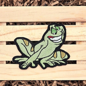 May include: Green embroidered frog patch with a toothy grin and yellow spots. The patch is on a wooden background.