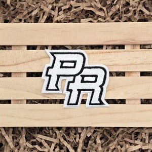 May include: White and black embroidered patch featuring the letters "PR" in a stylized font. The patch is rectangular and has a black border. The patch is placed on a light brown wooden crate with brown paper filler.