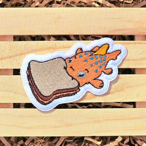 May include: An orange and blue cartoon fish patch with a sandwich. The fish has blue spots and a yellow fin. The sandwich has brown bread and a red and brown filling. The patch is outlined in white.