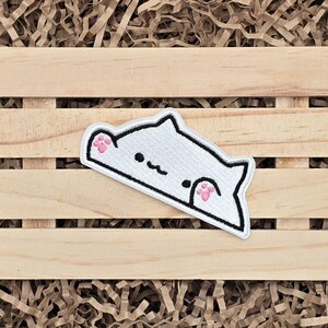 May include: White cat-shaped embroidered patch with black outline and pink paw pads. The cat has a simple, minimalist design with a smiling face. The patch is placed on a wooden surface with brown paper filler.
