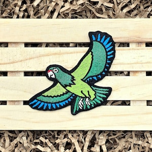 May include: Embroidered patch featuring a green parrot in flight with blue wing accents and a black border. The parrot has a red beak and white eye details. The patch is designed for application on clothing or accessories.