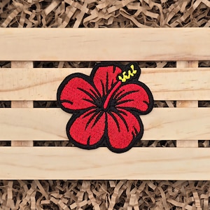 May include: Embroidered red hibiscus flower patch with black outline and yellow accents. The patch is placed on a wooden crate with brown paper filler. This floral patch is perfect for adding a tropical touch to clothing or accessories.
