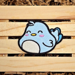 May include: A blue embroidered patch of a cartoon bird with a white belly and pink cheeks. The bird is smiling and has its wings outstretched.