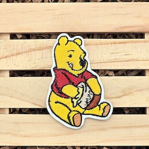 May include: Embroidered iron-on patch featuring Winnie the Pooh, a cartoon bear, in yellow and red, holding a honey pot.