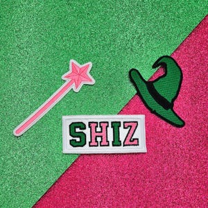 May include: A pink and green felt patch with the word "SHIZ" in white letters. The patch is on a green and pink glitter background. There is also a pink star wand and a green hat with a black brim.
