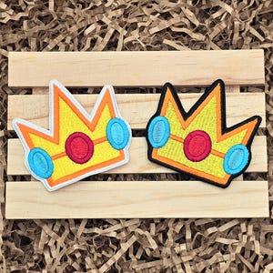 May include: Two embroidered crown patches. Each crown is yellow with orange trim, a red center, and two light blue side accents. One patch has a white border, the other has a black border. The patches are on a wooden pallet.