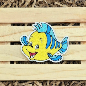 May include: Embroidered patch featuring a cartoon yellow fish with blue accents. The fish has a smiling expression and is outlined in white. The patch is likely for clothing or accessories.